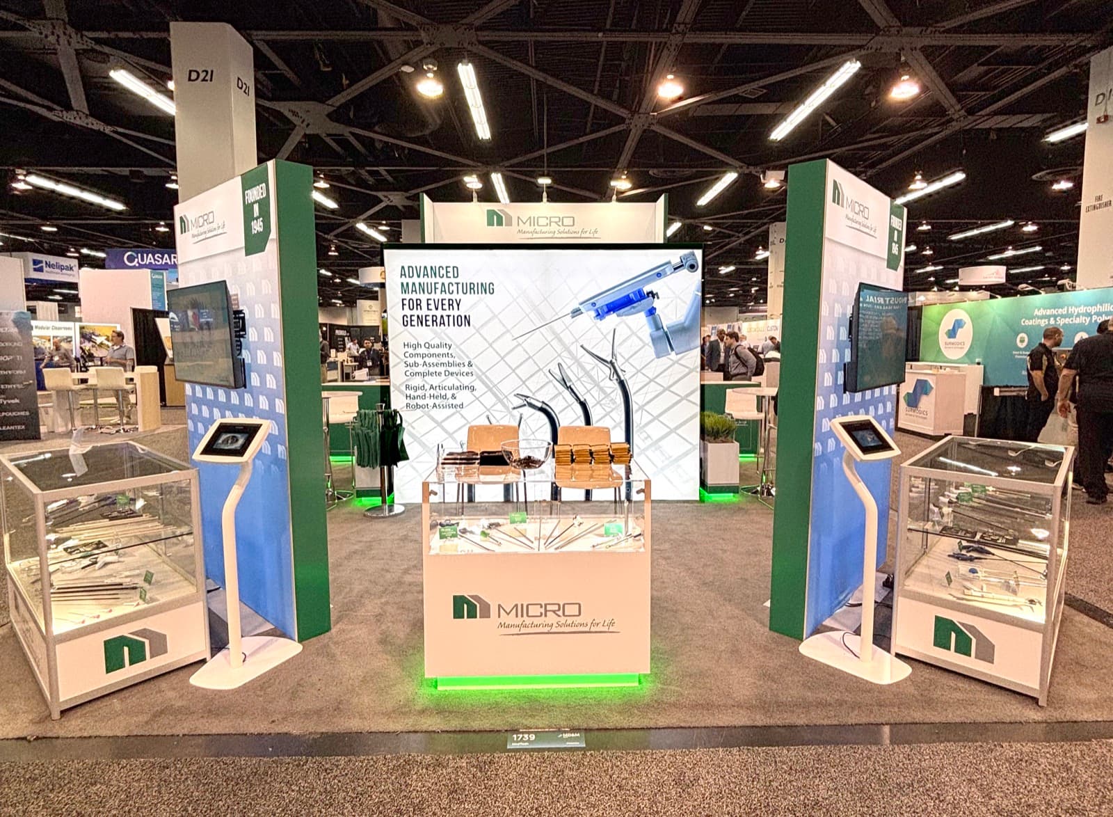 MICRO island trade show booth at MD&M with backlit tower graphics, glass display cases of surgical instruments, and interactive tablet kiosks, built and installed by HAP