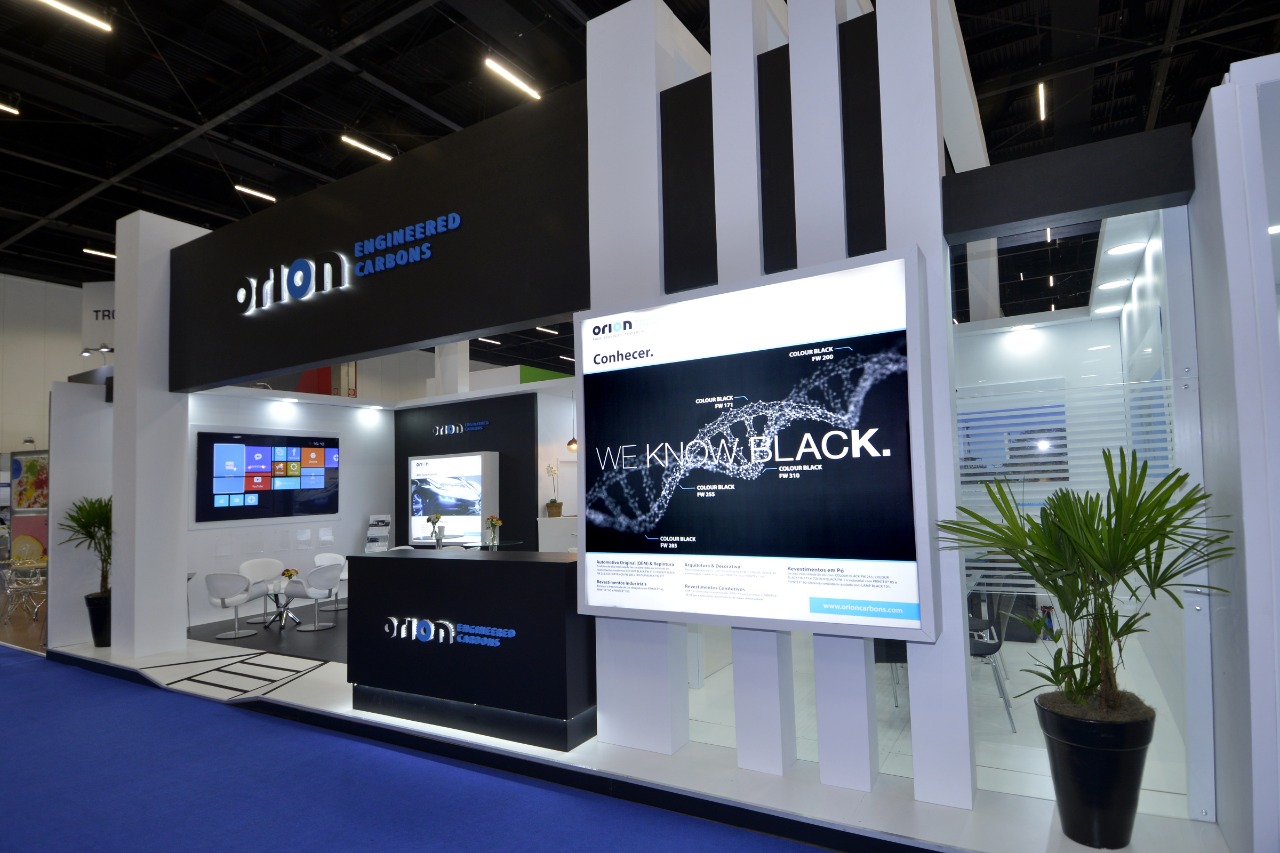 Black-and-white Orion Engineered Carbons trade show booth with illuminated logo fascia, backlit 'We Know Black' graphic wall, and lounge seating — designed and built by HAP