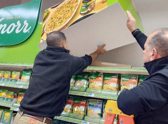 Two installers mounting a large Knorr display header above grocery shelves stocked with Knorr products